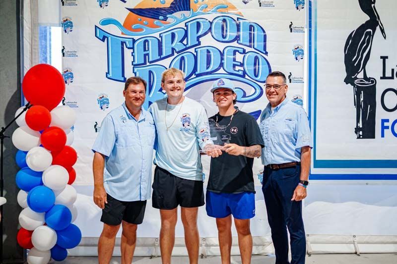 Four men posing with a trophy in front of a Tarpon Rodeo sign and red, white, and blue balloons