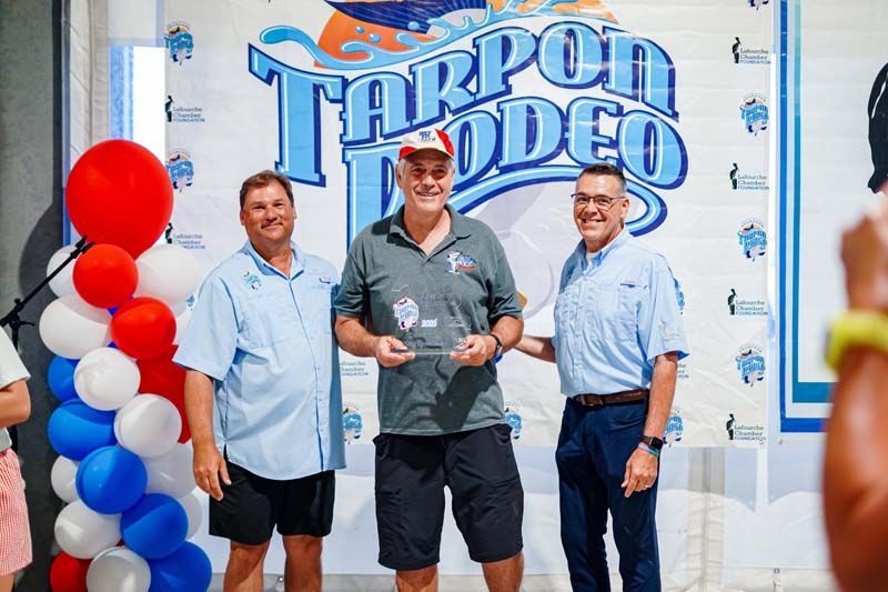 Three men stand at a Tarpon Rodeo event holding a trophy, with red, white, and blue balloons nearby.