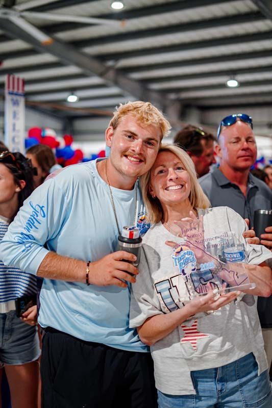 Two smiling people posing indoors at a crowded event, holding drinks and a souvenir tray.
