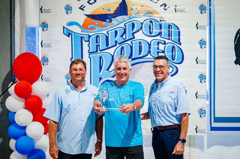 Three men pose in front of a Tarpon Rodeo banner, holding a trophy beside red, white, and blue balloons.