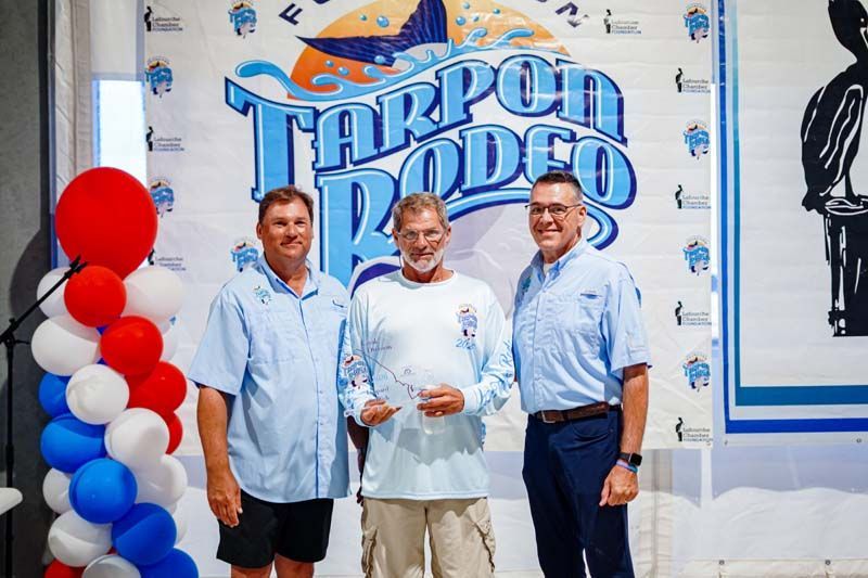 Three men posing in front of a Tarpon Rodeo sign with red, white, and blue balloons.