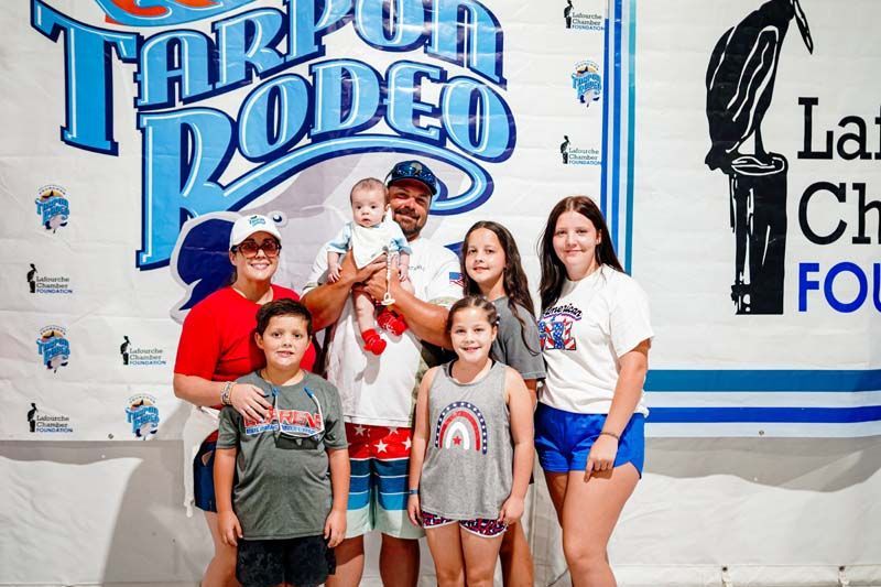 Group of children posing in front of a Tarpon Rodeo banner, some holding trophies and wearing event shirts