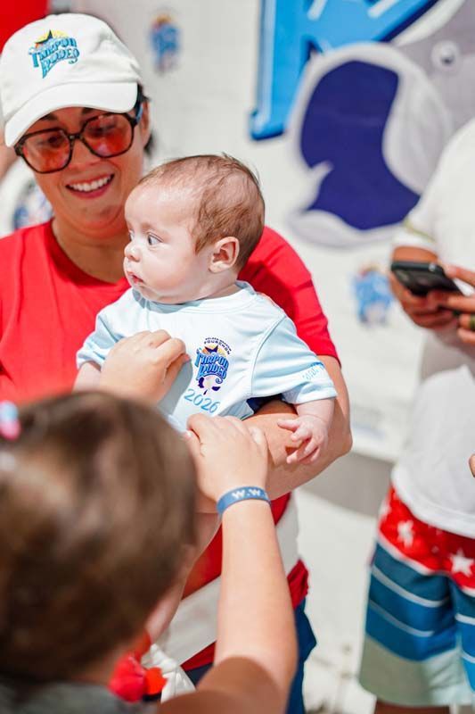 Woman in a white cap holding a baby at a colorful event, with people nearby
