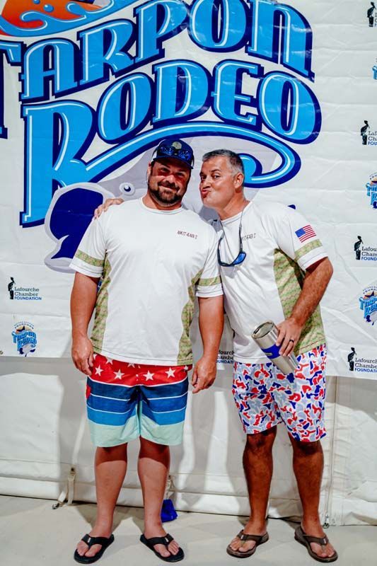 Two men standing in front of a Tarpon Rodeo sign, smiling in colorful beach shorts and flip-flops