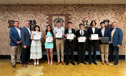 Group of students and adults posing with certificates in front of a brick wall at an awards ceremony.