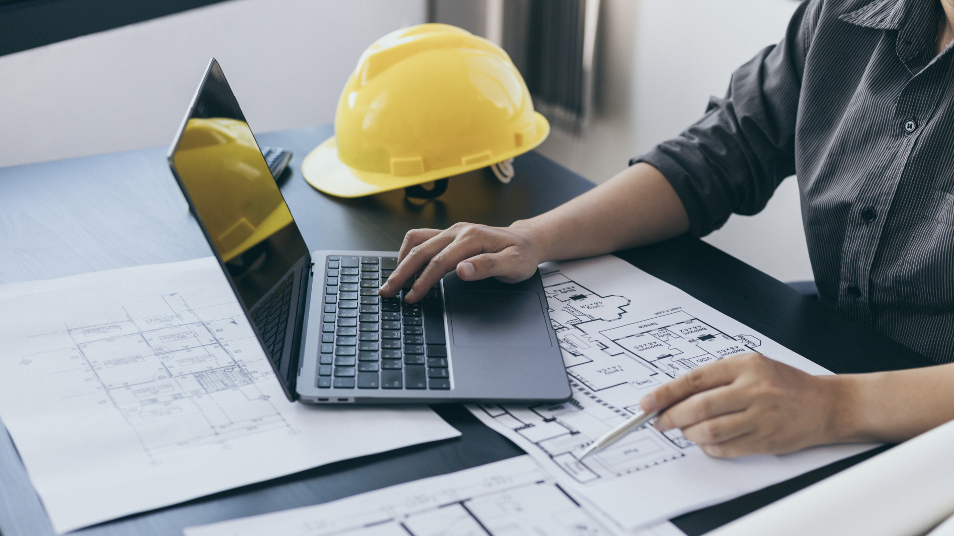 Engineer working on laptop over architectural blueprints, with a yellow hard hat nearby.