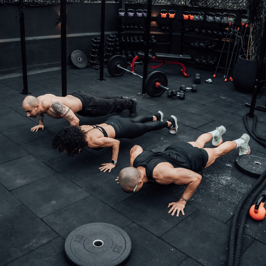 Four people doing push-ups in a dark gym with dumbbells and weight plates around them