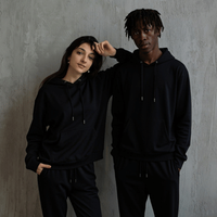 Two people in black hoodies and pants posing against a gray wall