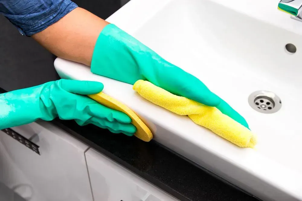 Hands in Green Gloves Cleaning a White Sink With a Yellow Sponge — Jamac Cleaning Services in Cardiff, NSW