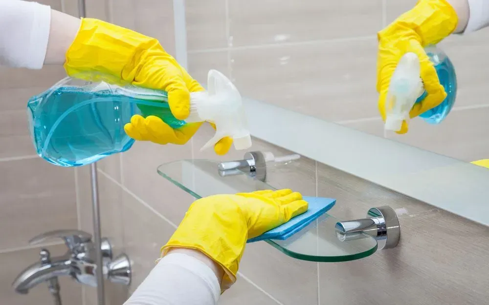 Hands in Yellow Gloves Cleaning a Bathroom Mirror With Spray and Cloth — Jamac Cleaning Services in Cardiff, NSW