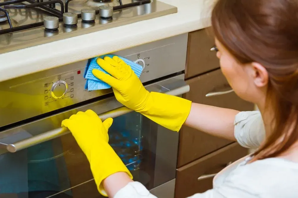 Woman Wearing Yellow Gloves Cleaning an Oven With a Blue Cloth in a Kitchen — Jamac Cleaning Services in Charlestown, NSW
