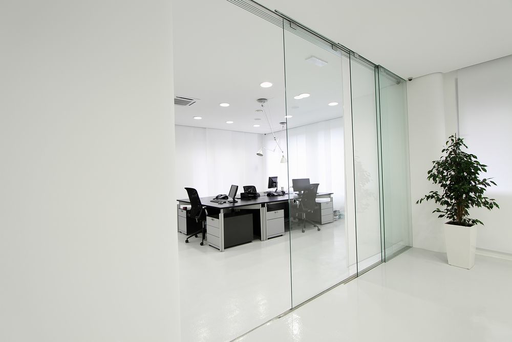 Minimal white office with glass walls, desks, chairs, and a potted tree in the corner.