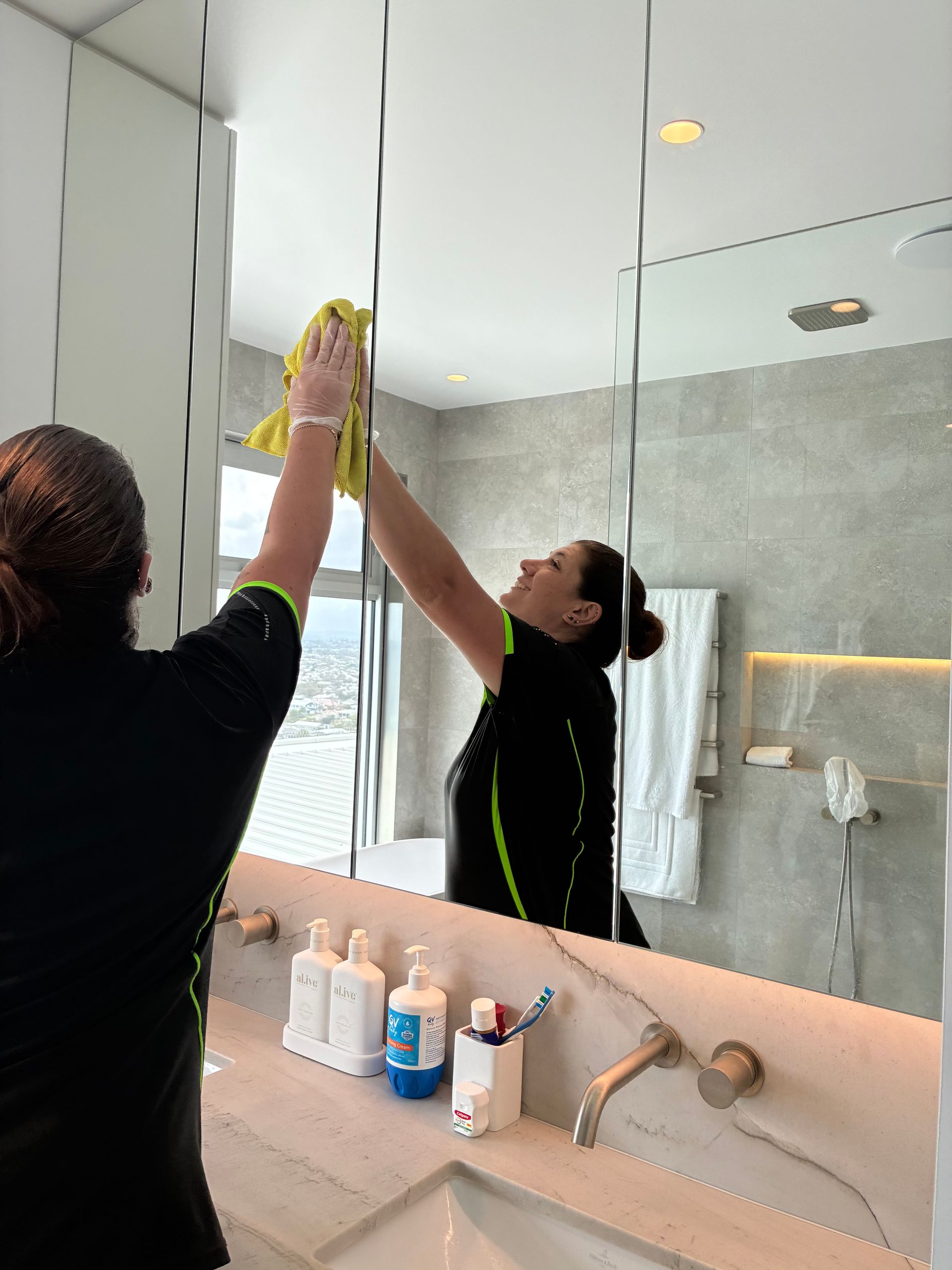 Person cleaning a bathroom mirror with a cloth, seen in the reflection above a sink.