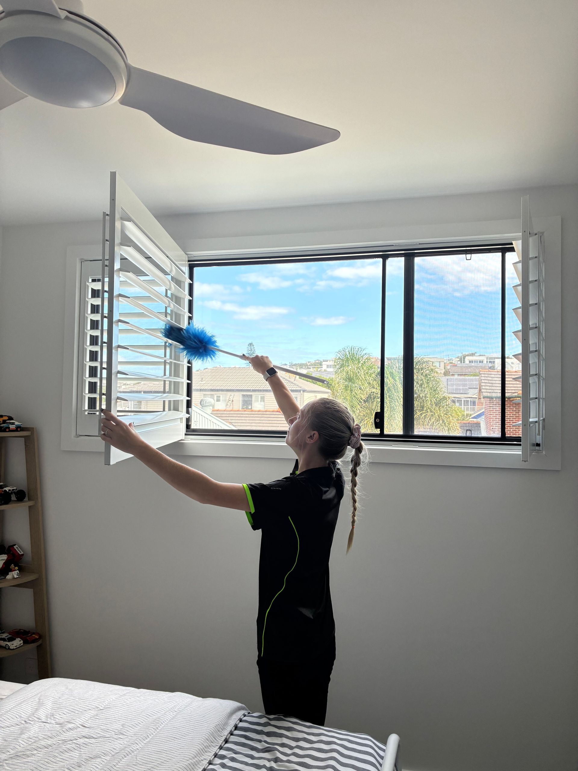 Child dusting open window shutters in a bright bedroom