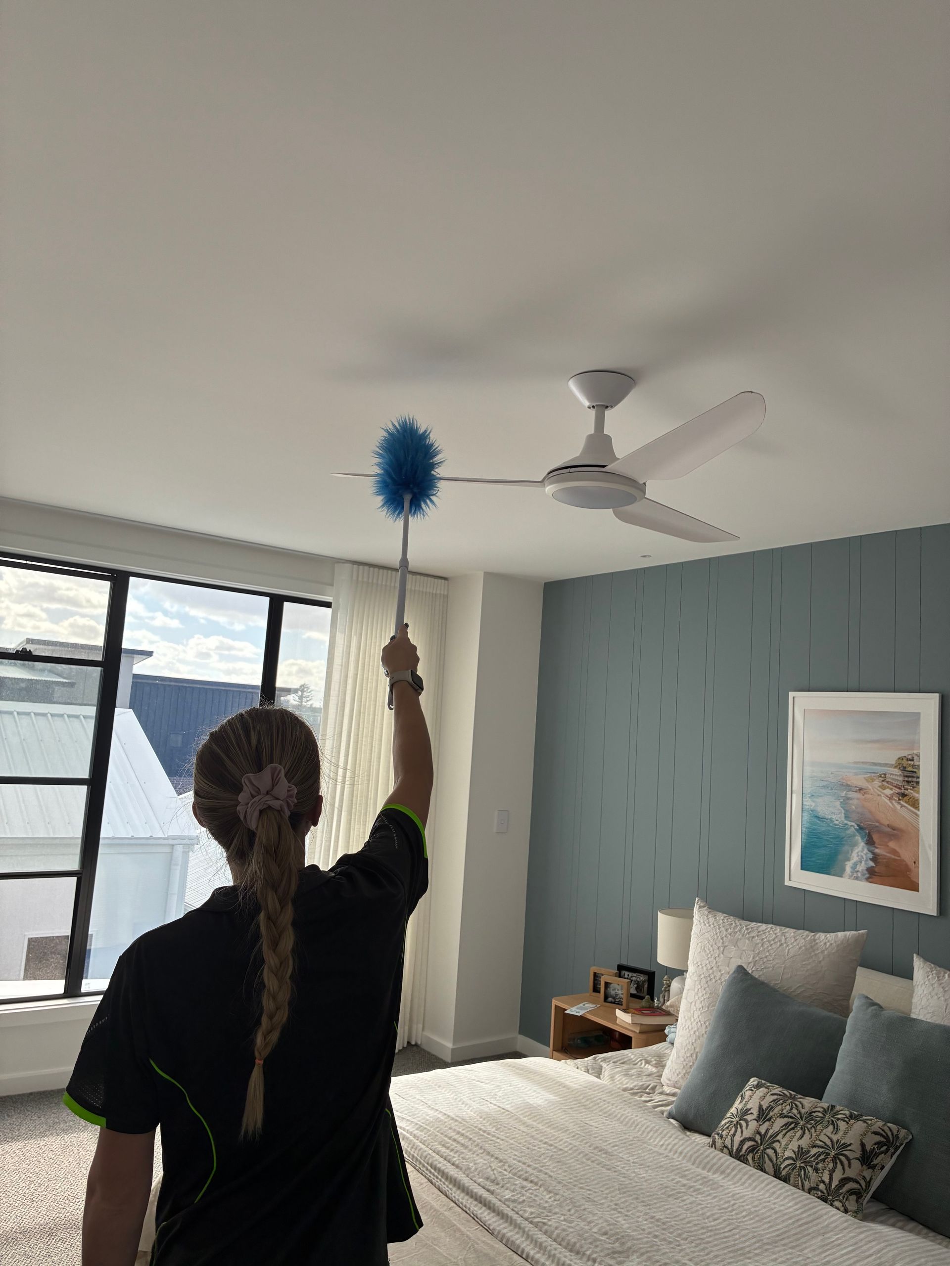 Person dusting a white ceiling fan in a bright bedroom with a bed, window, and mountain view.