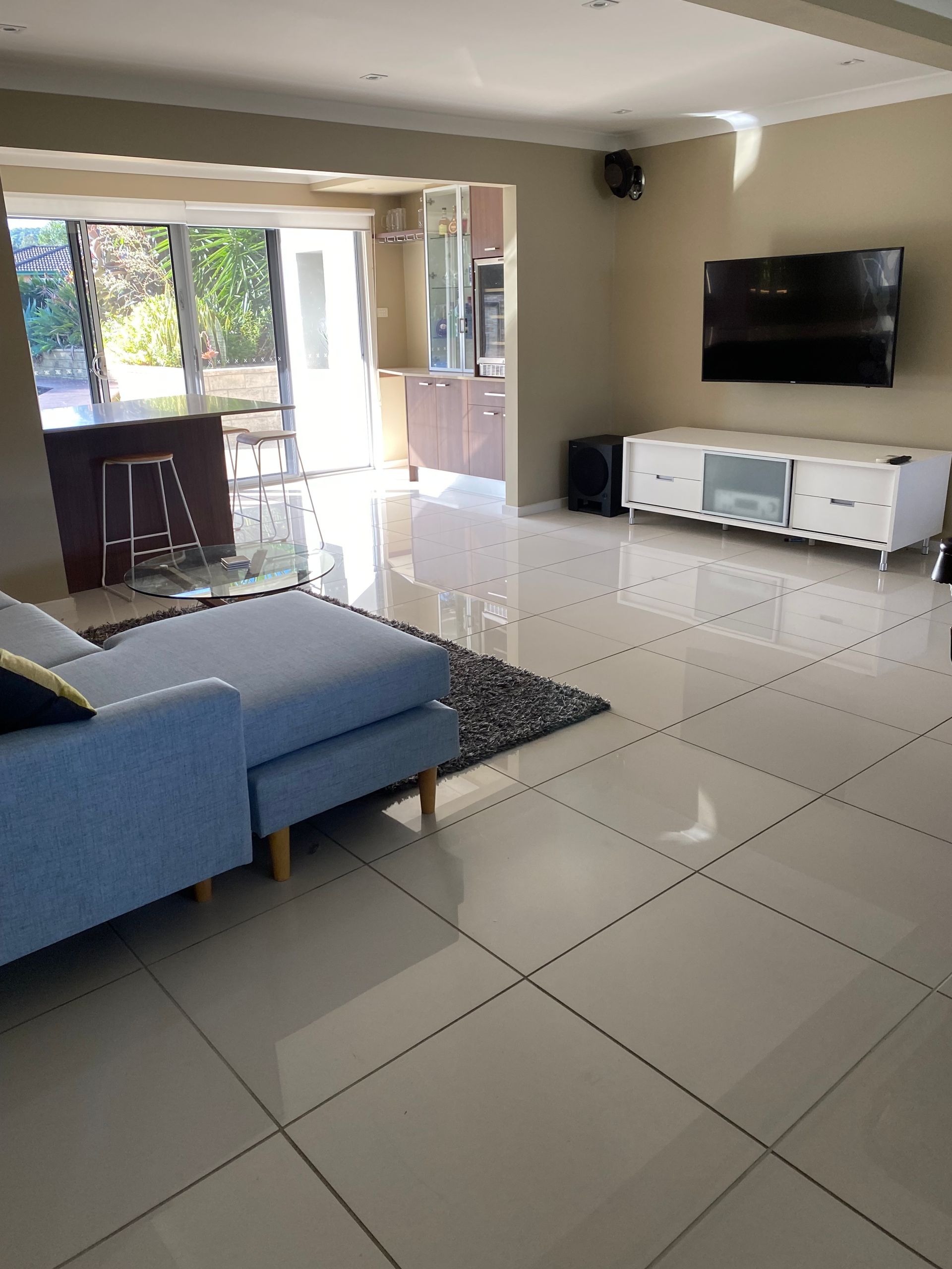 Bright living room with blue sectional, glossy tile floor, wall-mounted TV, and open doors to a patio