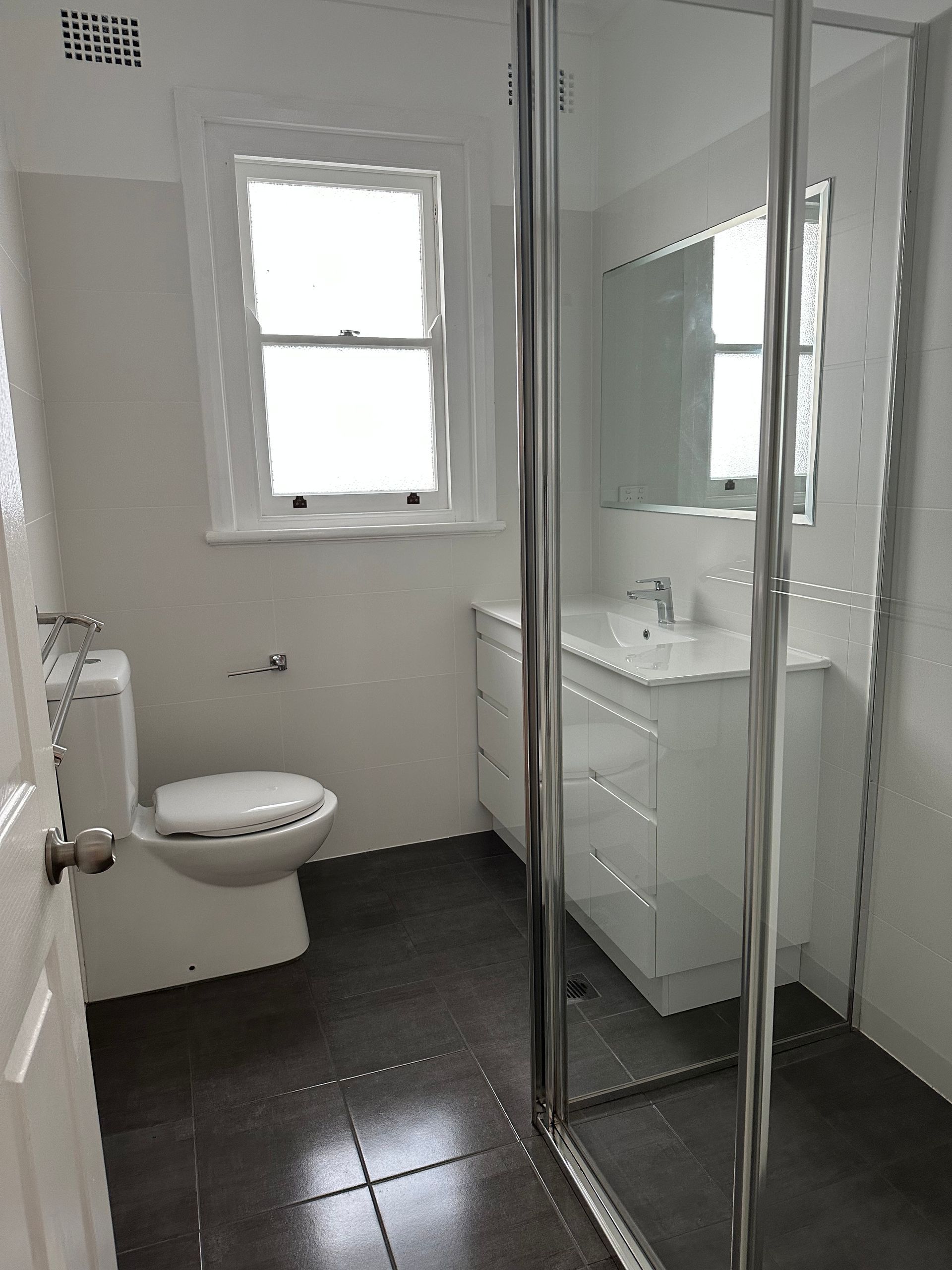 Small bathroom with toilet, window, sink vanity, and glass shower doors.