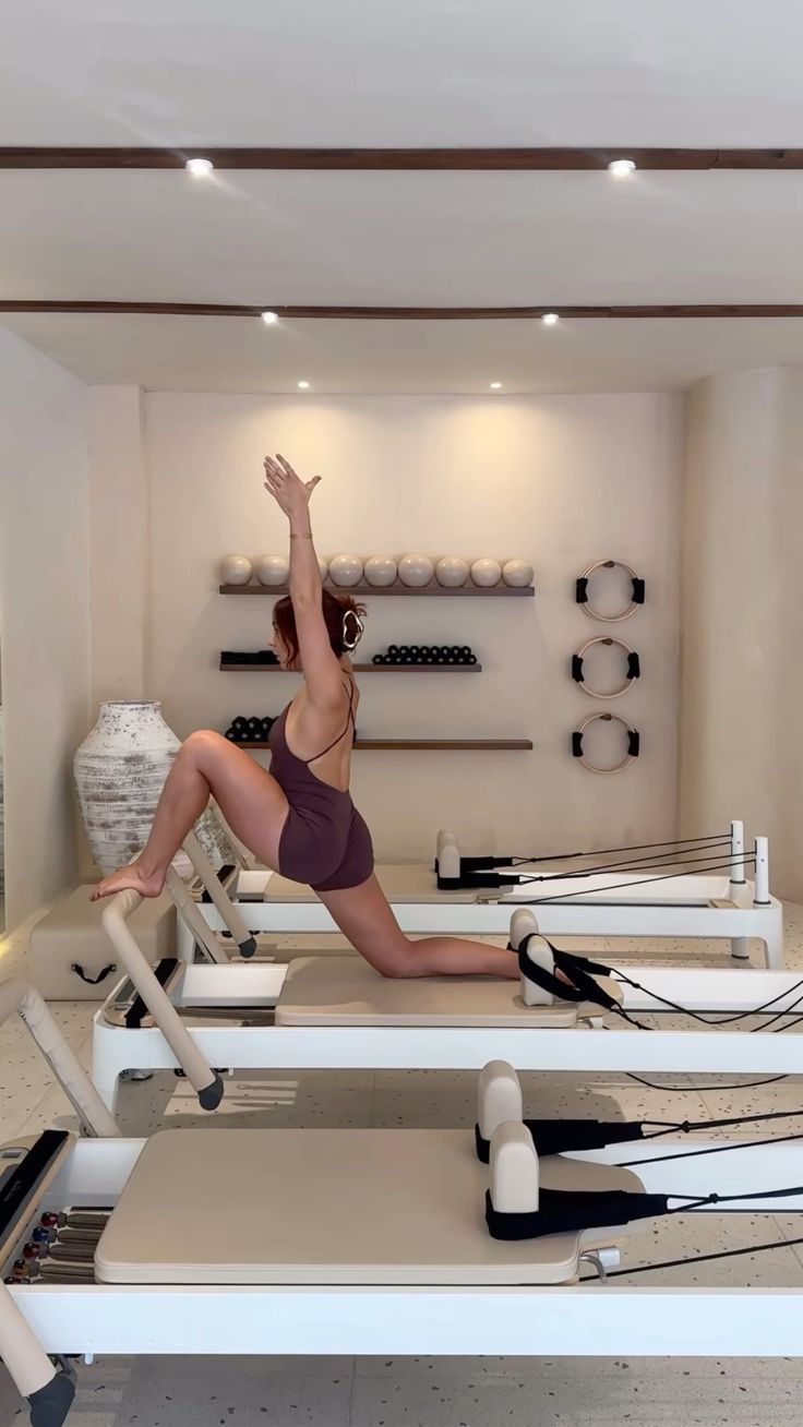 Woman doing Pilates on a reformer machine, kneeling with arms extended, light beige outfit in a studio.