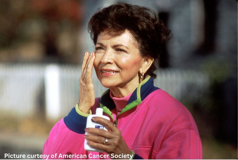A woman in a pink jacket is applying sunscreen to her face