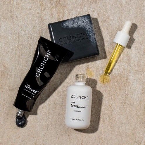 Four Crunchi skincare products on a textured beige background: a black tube, a soap bar, a white bottle, and a face oil.
