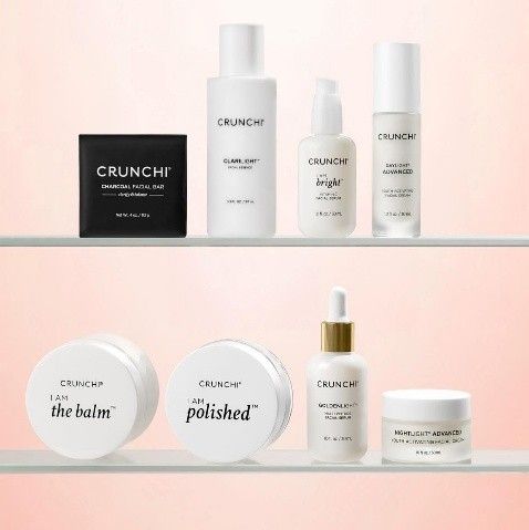 A collection of Crunchi skincare products arranged on two shelves against a soft, light pink background.