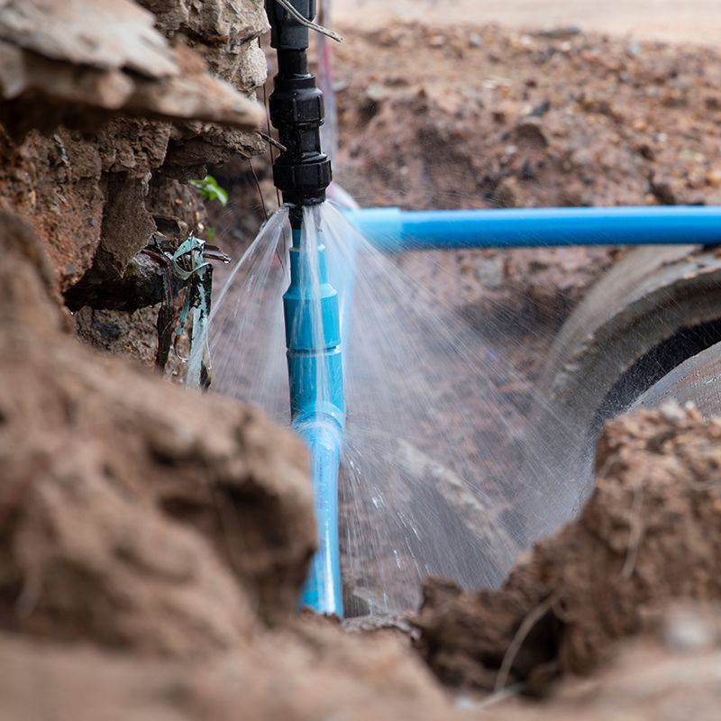 Leaking blue PVC pipe in a dirt excavation, spraying water.