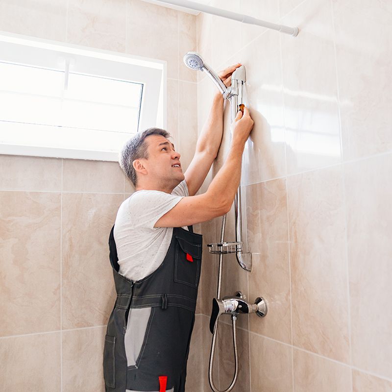 Man in overalls installing a showerhead in a tiled bathroom.