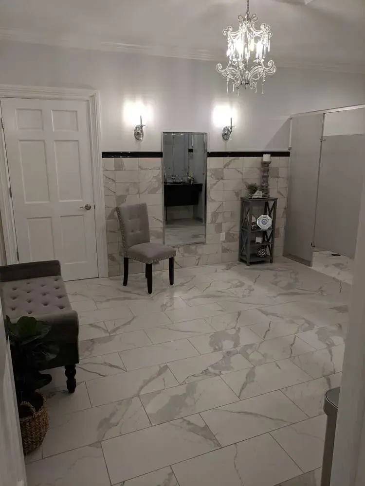 Bright marble-tiled room with chandelier, chairs, vanity table, and open doorway to a bathroom.