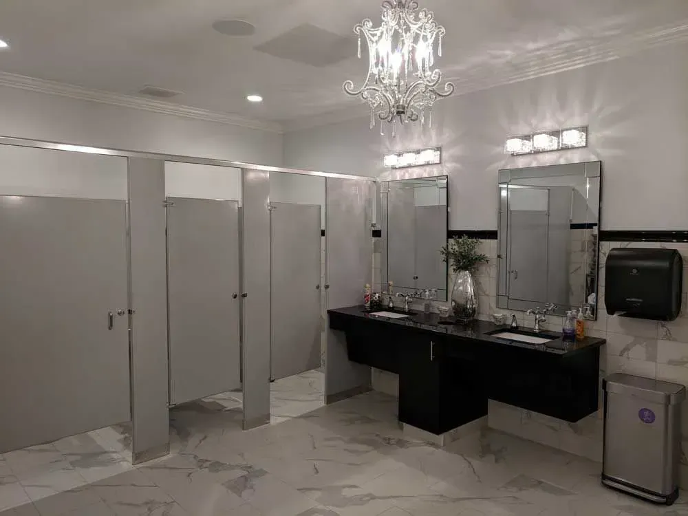 Bright restroom with white stall doors, black double sink vanity, marble floor, and chandelier lighting
