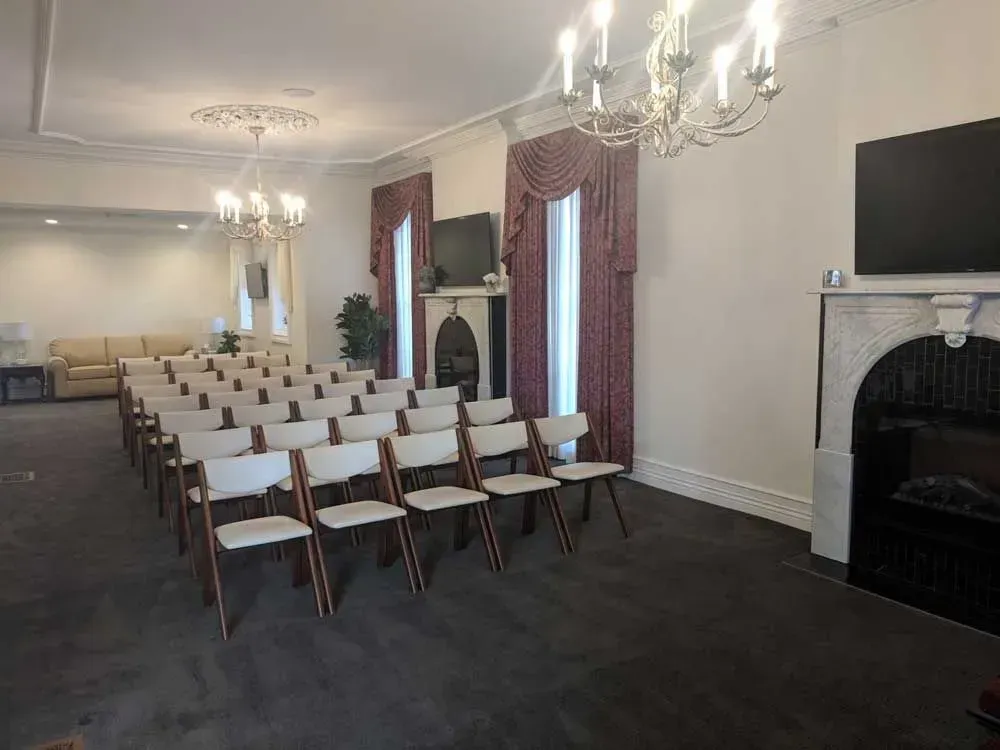 Empty event room with rows of white chairs, chandeliers, carpeted floor, and a fireplace and TV at the front
