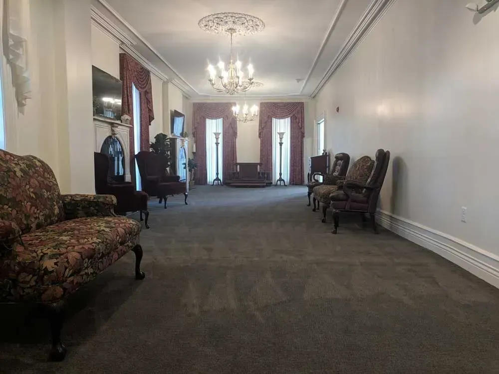 Elegant hallway with chandeliers, floral armchairs, and tall windows along a carpeted floor.