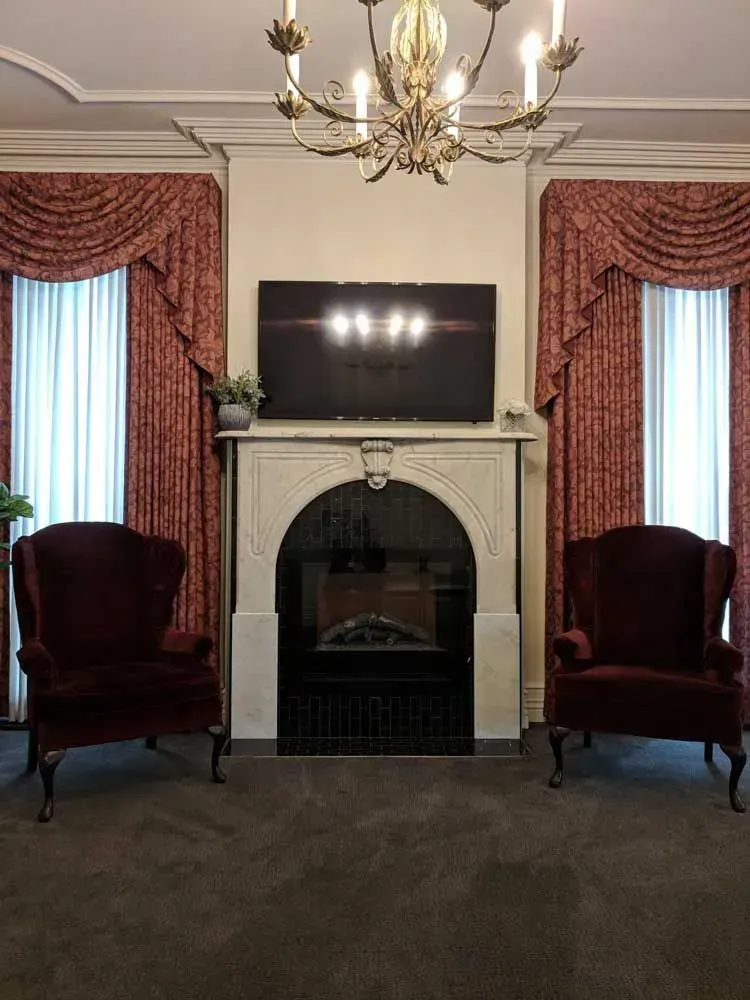 Elegant room with fireplace, TV, chandelier, and two burgundy velvet chairs by tall windows.