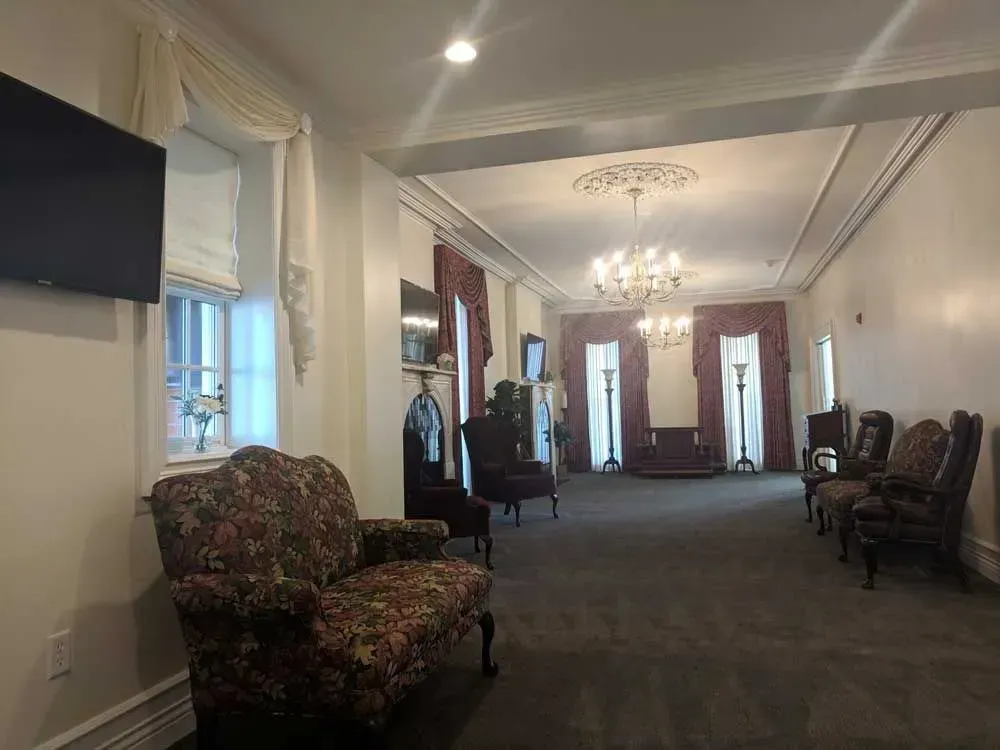 Elegant hallway with chandelier, dark carpet, floral armchair, and row of chairs along the walls.