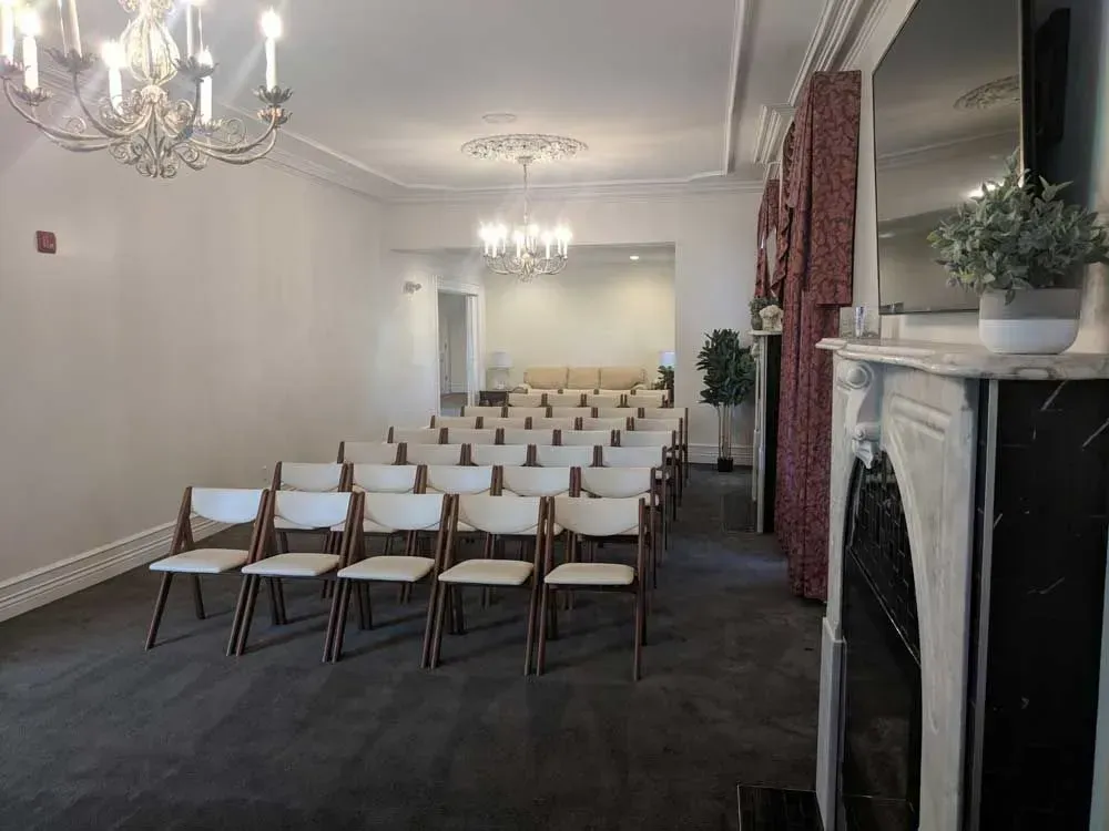 Empty white event room with rows of chairs, chandeliers, and a fireplace mantel