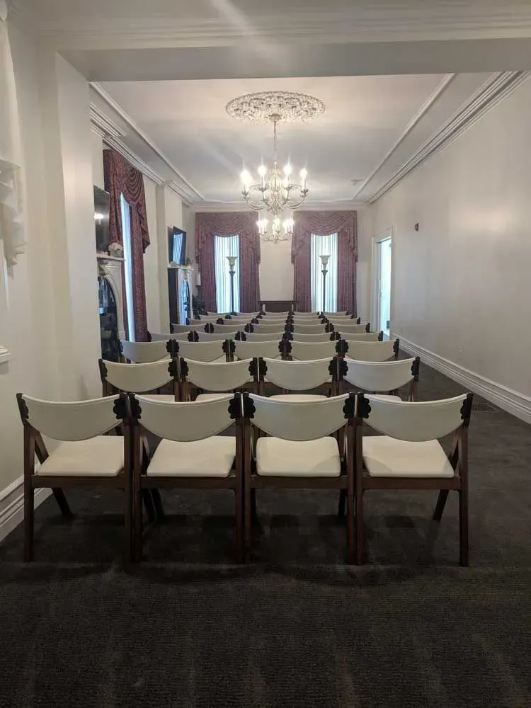 Rows of white chairs facing a chandelier in a long, elegant room with tall windows and dark carpet