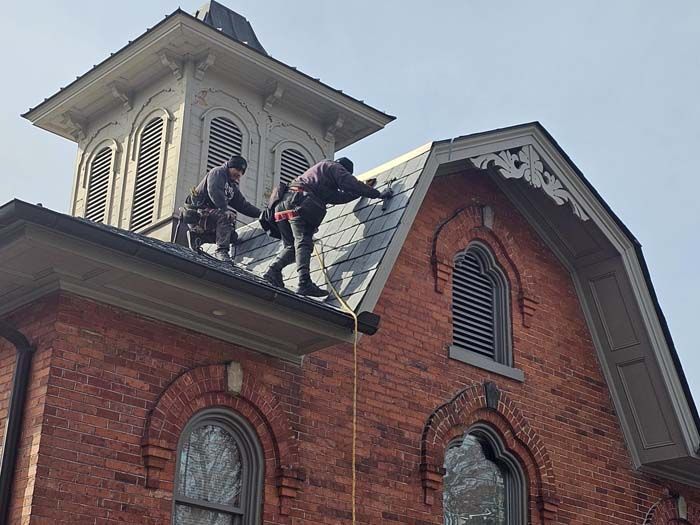 Two workers on a brick church roof repairing shingles near a white steeple