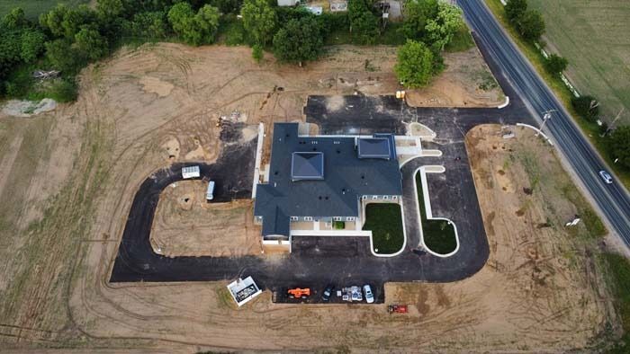 Aerial view of a house under construction with dirt lot, paved driveway, and a road beside trees.