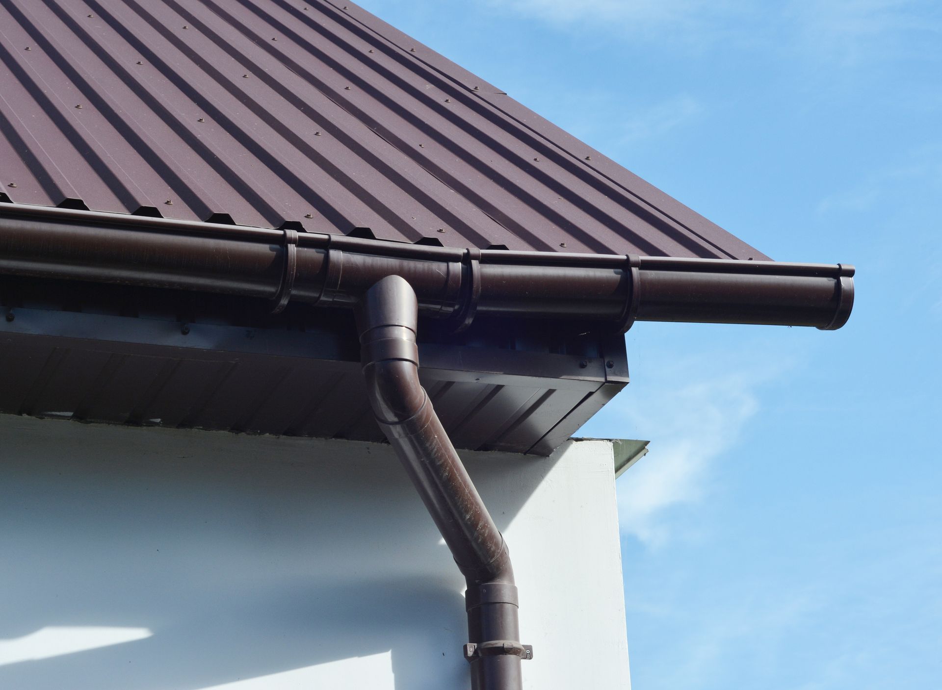 Brown metal roof with fascia, soffit, and rain gutter on a white wall under the sky.