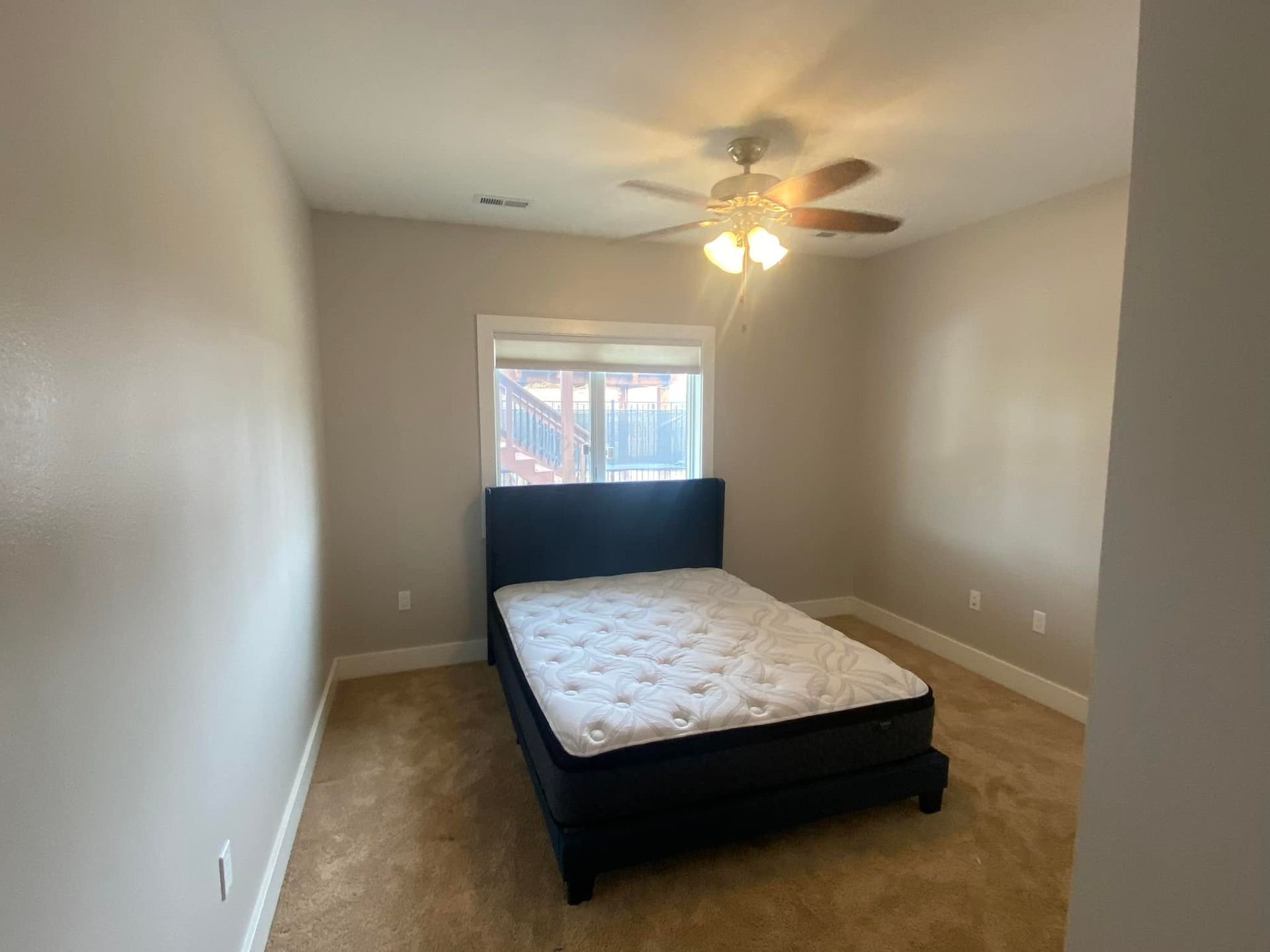 Bedroom with bed, window, ceiling fan, and beige walls and carpet.