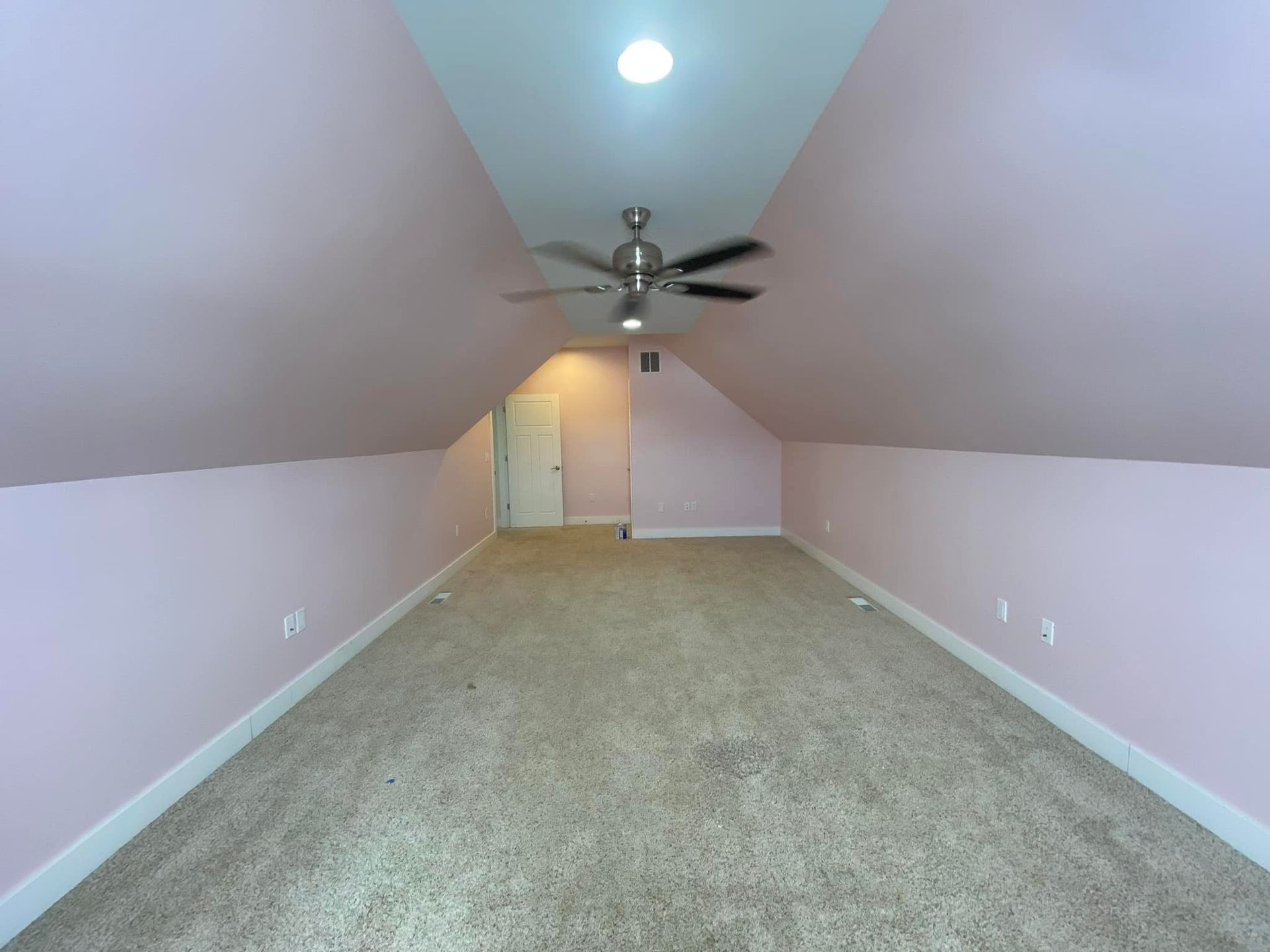 Empty attic room with pink walls, tan carpet, and a ceiling fan.
