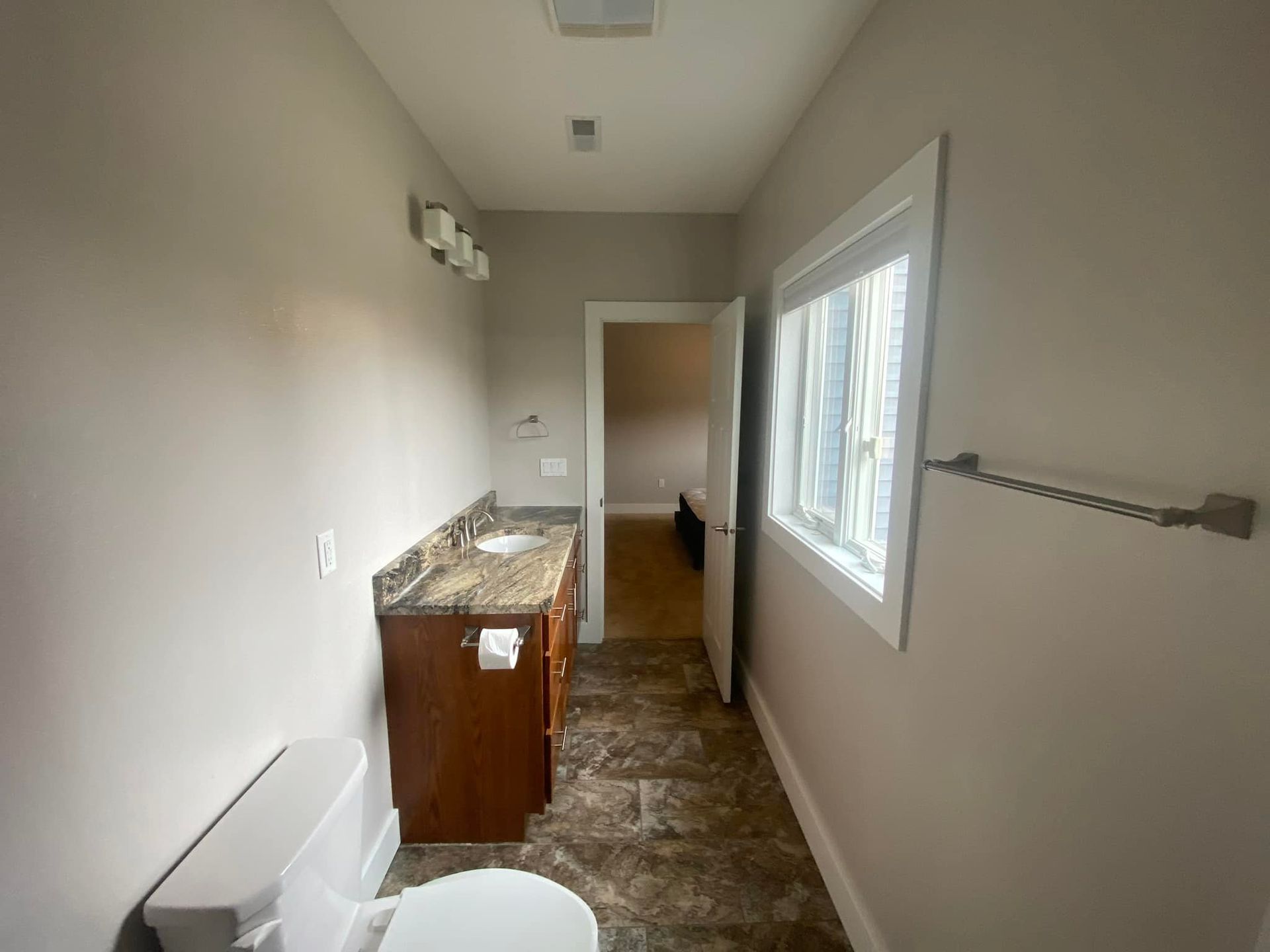Bathroom with vanity, toilet, window, and doorway to a bedroom. Light brown walls, dark brown cabinets, and tile floor.