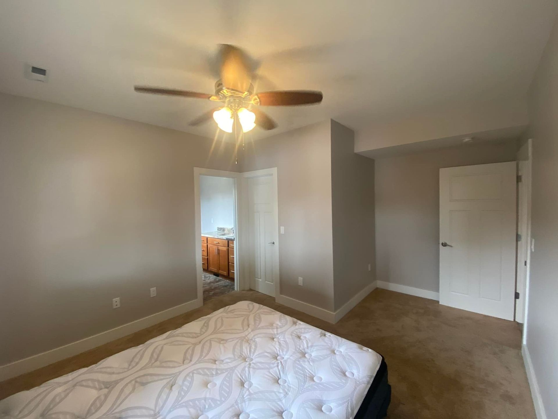 Bedroom with tan walls, beige carpet, bed, ceiling fan, and a door leading to a bathroom.