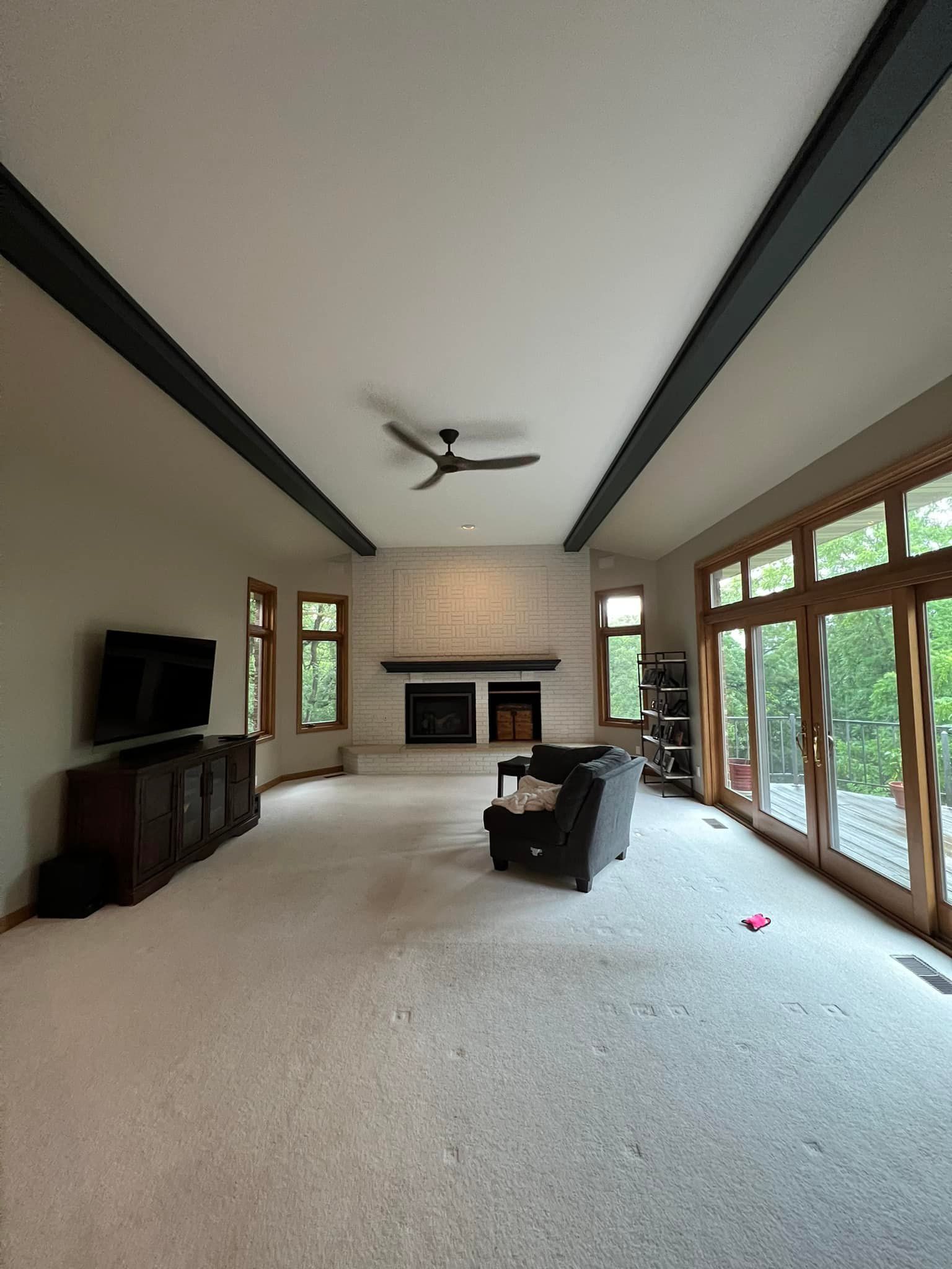 Spacious living room with gray walls, beige carpet, dark beams, fireplace, TV, and large windows.