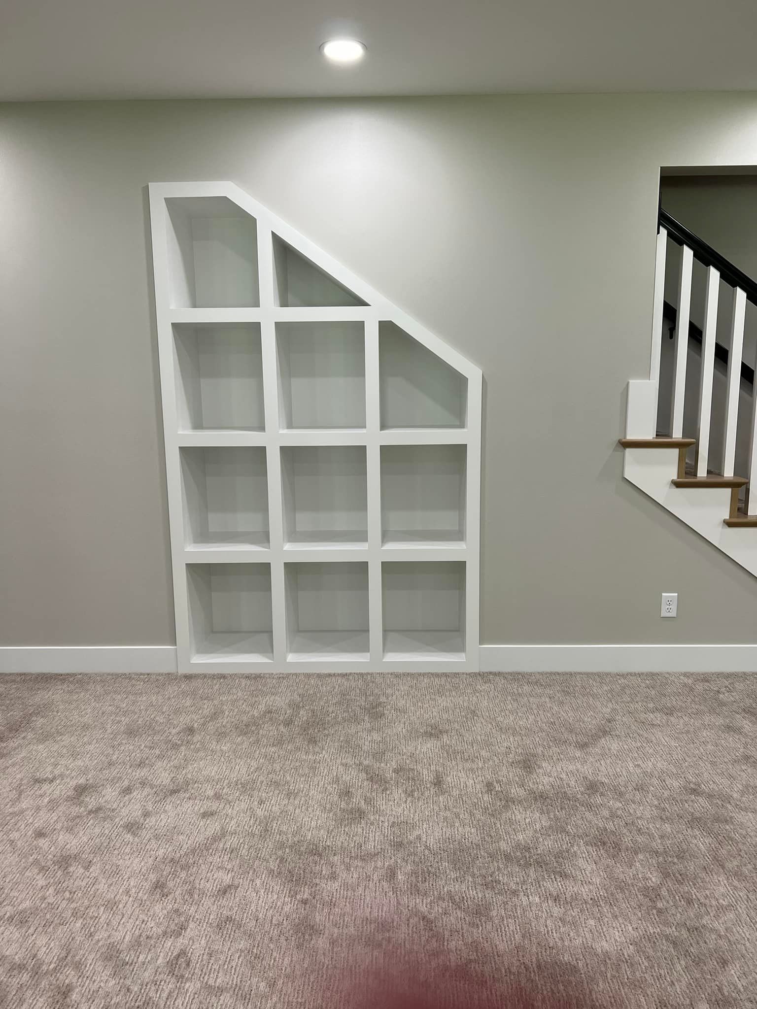 White built-in shelving unit with angled top on a gray wall next to a staircase. Carpeted floor.