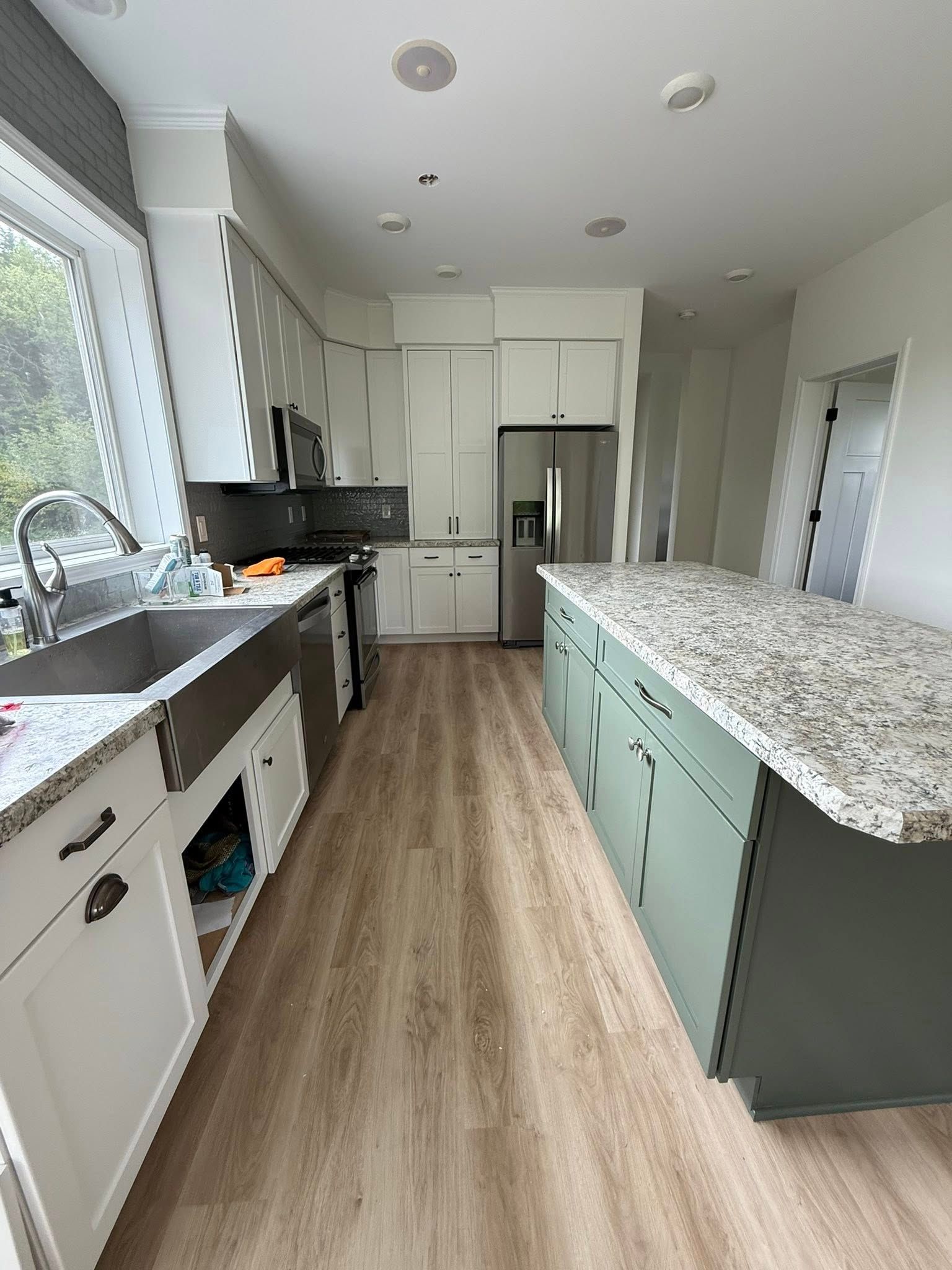Kitchen with green island, stainless steel sink, white cabinets, light wood-look flooring, and a window.