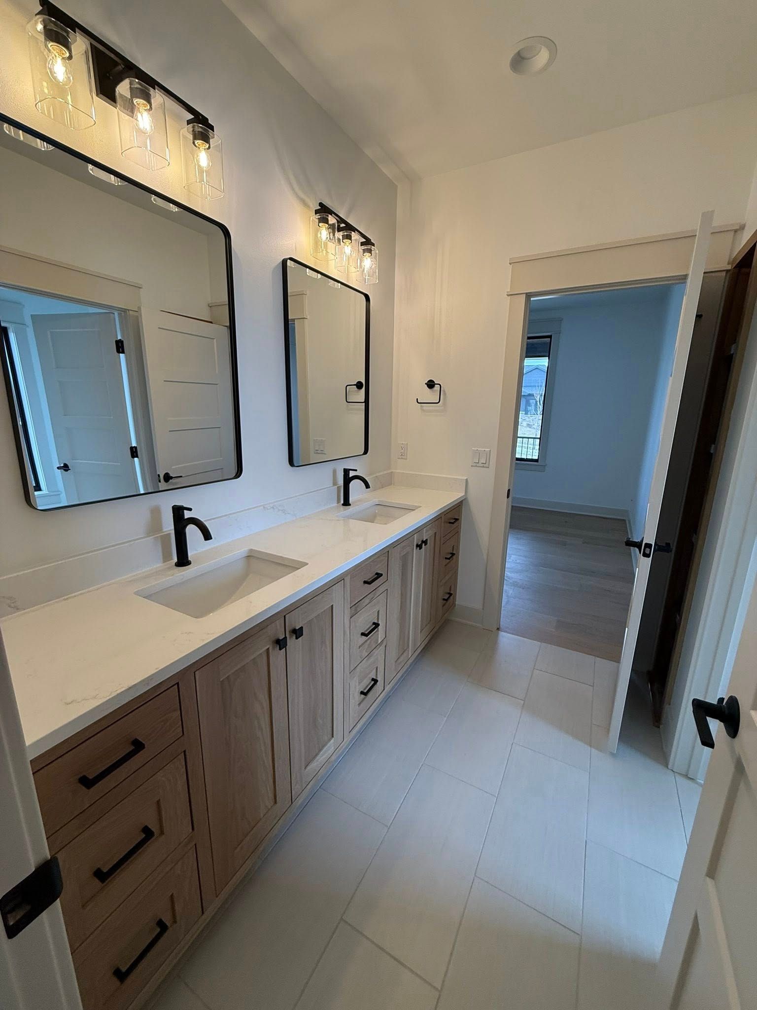 Bathroom with dual sinks, mirrors, wooden cabinets, black fixtures, and a doorway to another room.