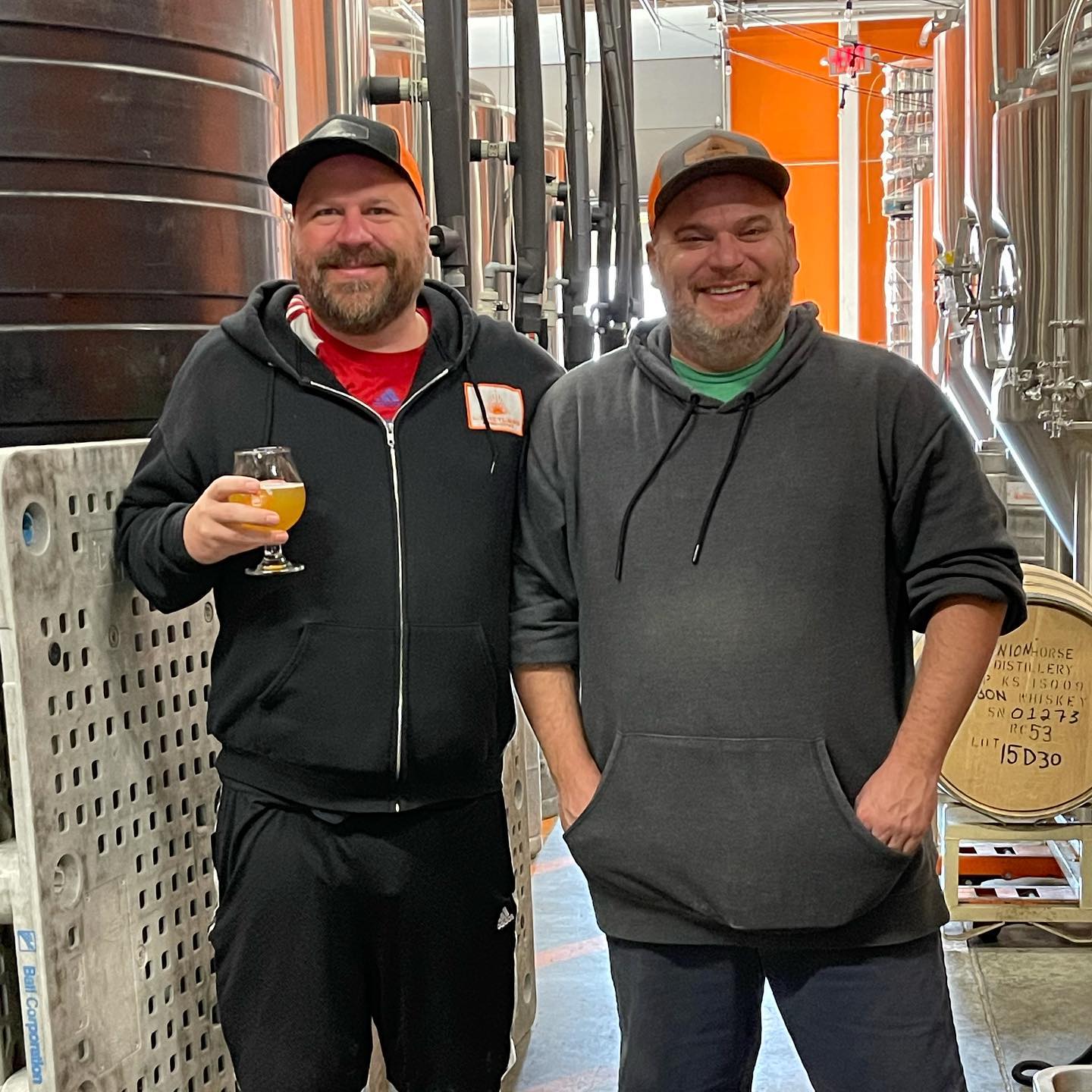 Two men standing in a brewery, one holding a beer, with stainless steel tanks in the background.