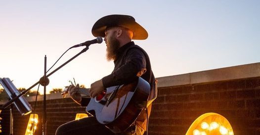 Musician in a cowboy hat playing guitar and singing on an outdoor stage at sunset