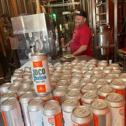 Man behind a table stacked with cans of Taco Joes beer in a brewery setting