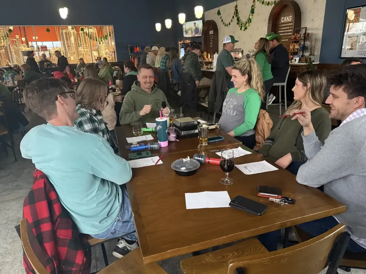 People chatting around a wooden table in a lively bar or café, with drinks and plates on the table.